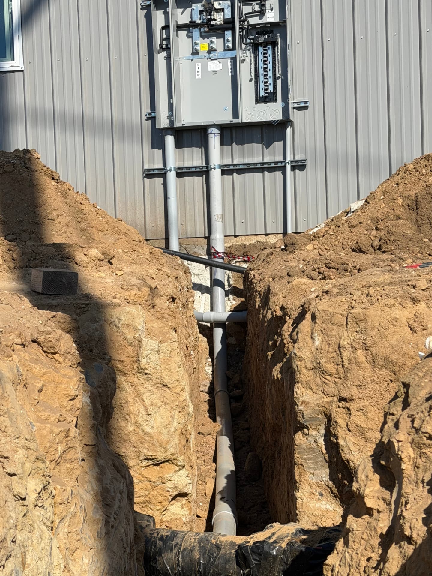 Open wall-mounted electrical service enclosure with vertical conduits extending into an excavated trench